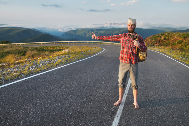 一个留着胡子的年轻嬉皮士，戴着衬衫帽子，赤脚走在山间的乡间小路上。搭便车和免费旅行图片下载