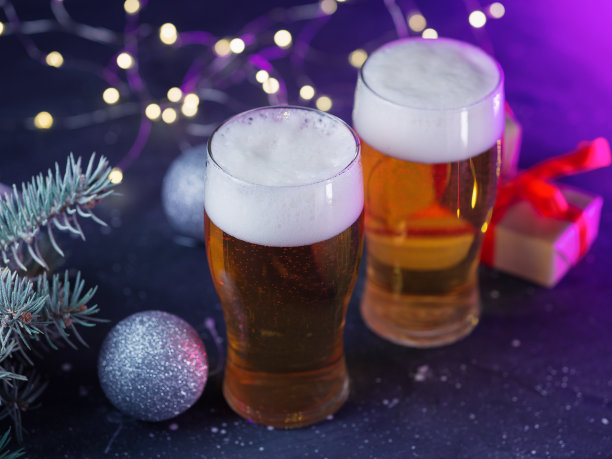 两杯淡啤酒放在装饰过的节日桌子上，上面摆满了礼物、花环和树枝。彩色光背景图片下载