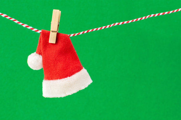 圣诞老人红白相间的皮帽上挂着一根绳子，上面绑着一个木制的晒衣夹。传统象征新年的服装以绿色为背景，复制空间。图片下载
