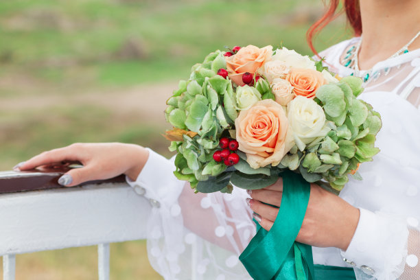 在婚礼当天，新娘手持一束由米色、橙色玫瑰和绿色树叶组成的美丽的婚礼花束。素材图片
