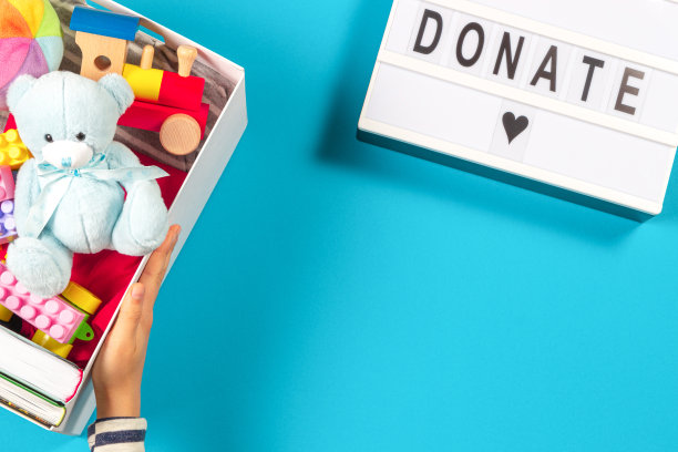 手拿捐赠箱与玩具、书籍、服装和灯箱文字捐赠浅蓝色背景。俯视图图片下载