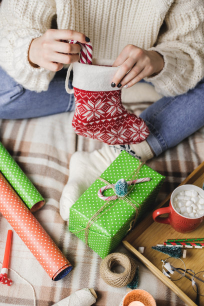 年轻的女人穿着舒适的针织羊毛衫坐在格子上，把条纹棒棒糖放在圣诞袜里作为礼物。准备寒假。木制托盘，一马克杯可可和棉花糖。图片下载