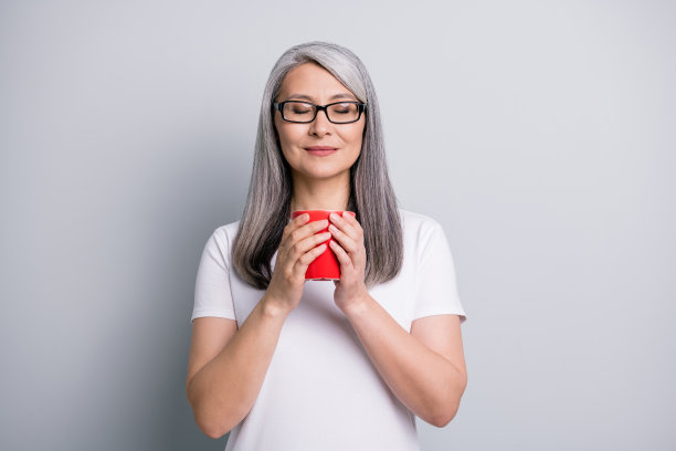 照片上的漂亮的老女人穿着休闲的衣服，戴着眼镜，手，胳膊，杯子享受香气，闭着眼睛孤立的灰色背景图片下载