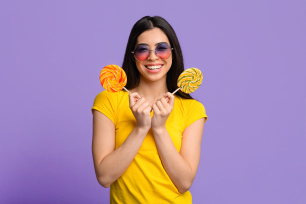 香甜可口。漂亮年轻的夏天女士用大明亮的棒棒糖遮住眼睛，看着相机，黄色的背景与拷贝空间图片下载