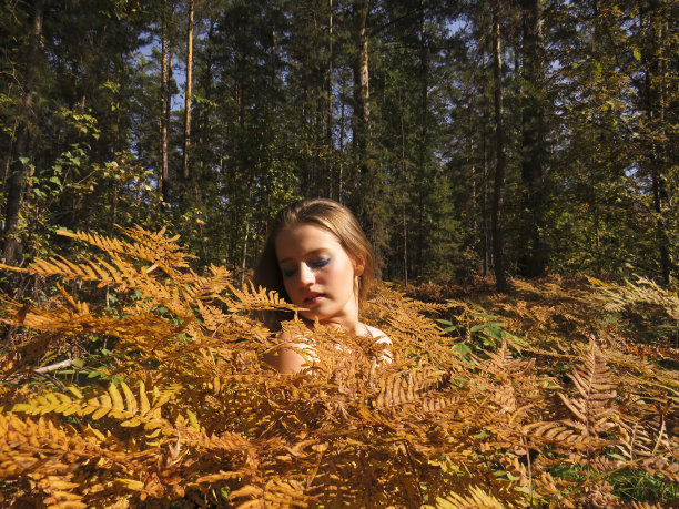 一个年轻的金发女人赤裸着肩膀，在秋天的森林里的一丛黄色蕨类植物中图片下载