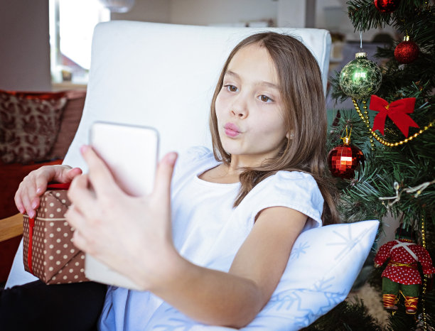 近距离微笑的高加索年轻女孩打电话，坐在附近的节日圣诞树在家里，快乐的女性拿着智能手机礼物盒，与朋友或亲戚聊天图片下载