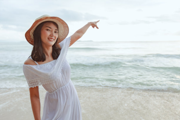 亚洲女人戴着时髦的帽子和衣服指着天空，看着大海，在海滩上享受美丽的日落。图片下载