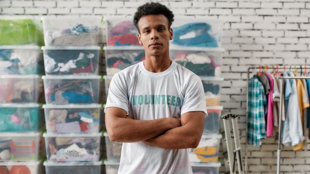 一个开朗的男人看着镜头，双臂交叉站在装满衣服的架子和箱子前。年轻的男性志愿者穿着t恤为慈善机构工作，为有需要的人捐赠衣服图片下载