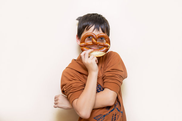 一个十几岁的男孩手里拿着一个开胃的巴伐利亚椒盐卷饼。用椒盐卷饼当面具看着摄像机。好玩的心情。面包店的概念图图片下载