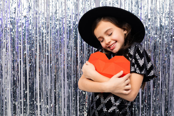 可爱的女孩微笑着，穿着节日的服装，戴着帽子，在闪闪发光的背景上抱着一个红色的纸心。情人节图片下载