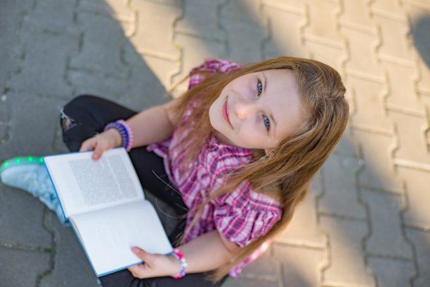 微笑的小女孩坐在人行道上看书。图片下载