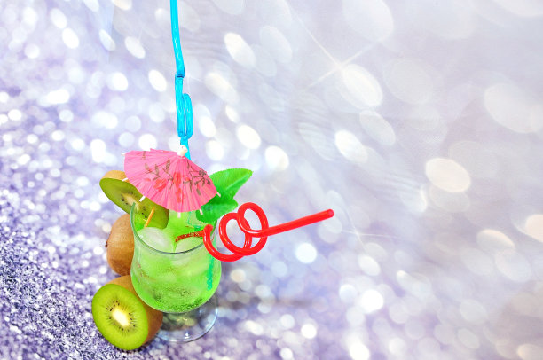 一杯加冰的绿色饮料，用鸡尾酒伞和吸管装饰在灰色的抽象背景上，旁边是成熟的猕猴桃。图片下载
