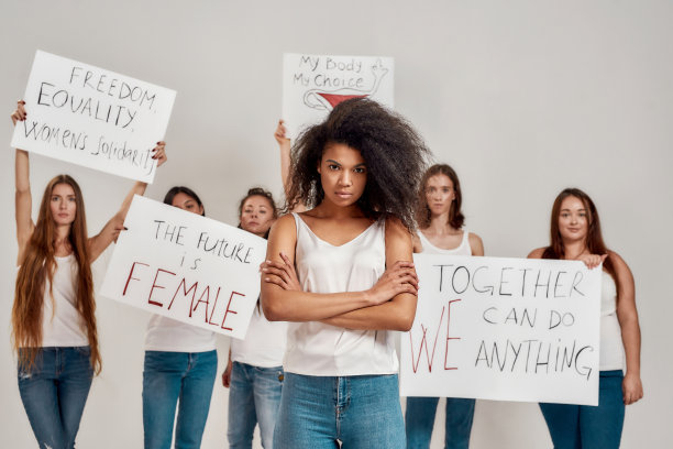 年轻的非洲裔美国妇女在白衬衫看着镜头。在背景中，一群不同的妇女高举着争取妇女权利的抗议横幅图片下载