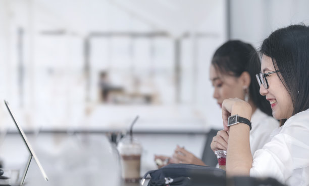 年轻的亚洲女人戴着眼镜微笑与幸福，而工作与电脑在现代办公室与她的朋友。图片下载