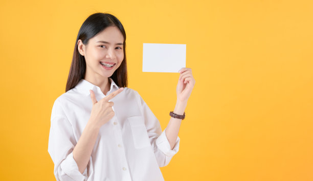 年轻的亚洲妇女拿着空白纸，微笑着看着黄色的背景。广告的迹象。图片下载