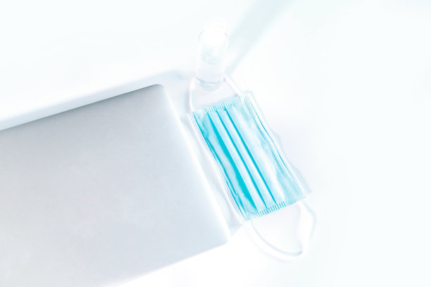 配有医用口罩和洗手液的现代笔记本电脑放在一张白色的桌子上。图片下载