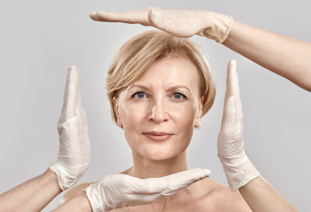 有吸引力的中年妇女看着相机的肖像。美容师创建一个框架周围女性的脸与手套的手孤立在灰色背景图片下载