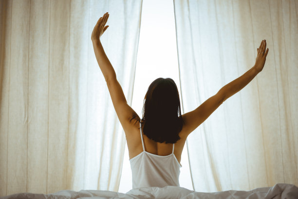 女人在床上伸开双手醒来后，太阳耀斑。深色头发的女人进入一个愉快和放松的一天后，睡了一个好觉和后视图。崭新的一天和快乐的周末图片下载