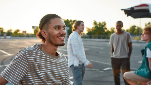 一个穿着随意、皮肤黝黑的拉丁裔男子的特写，他带着迷人的微笑站在停车场外面，背景是他的朋友图片下载