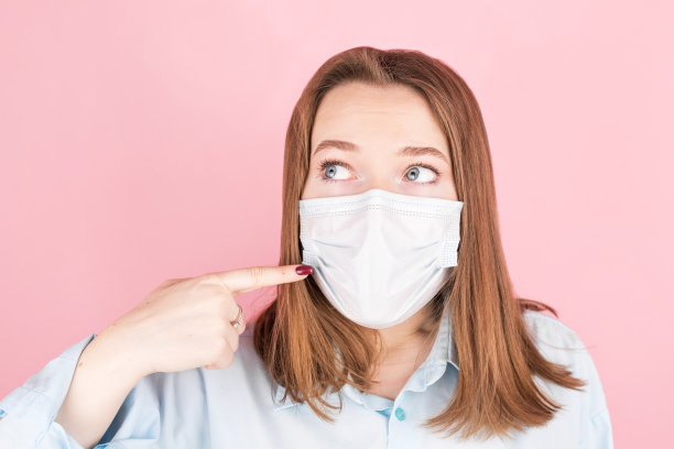 年轻迷人的女人用手指指着她脸上的医用防护面具。预防冠状病毒感染和感冒。图片下载
