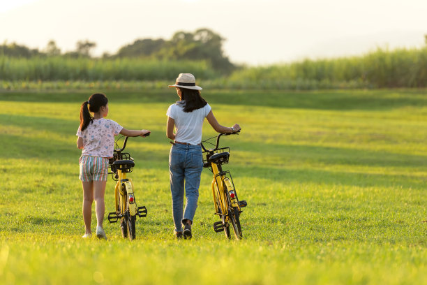 母亲和女儿骑着自行车在花园草地在日落附近的白色篱笆。家庭生活方式的概念图片下载
