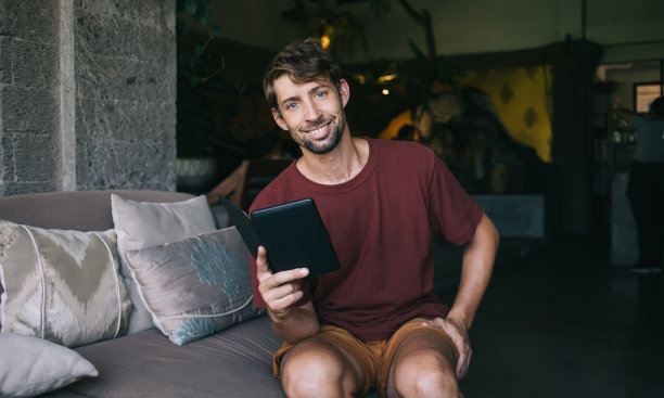 这是一幅穿着休闲装的开朗的白人男性看着相机，拿着电子阅读器在业余爱好上消磨时间，微笑着的时髦男学生用现代设备和4G享受教育的照片图片下载