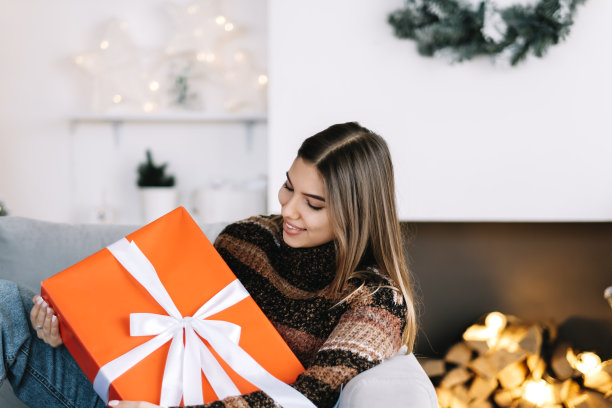 快乐的女人拿着大圣诞礼物在家里的沙发上的圣诞树附近。素材图片