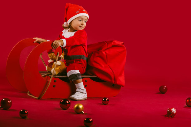 一个可爱的小女孩穿着红色的圣诞老人服装，坐在雪橇上，驯鹿带着礼物和冬天的装饰品。红色背景上的冬季概念。节日快乐图片下载