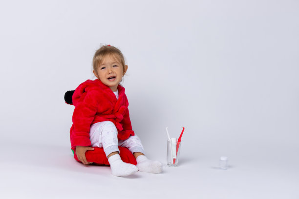 一个穿着睡衣和红袍子，拿着牙科工具箱的女孩坐在枕头上，哭着看着镜头。白色背景。图片下载