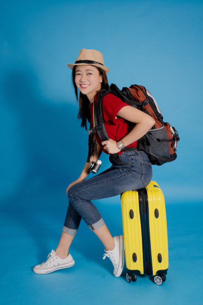 亚洲女游客在休闲衣服微笑和坐在手提箱孤立的蓝色背景。她喜欢周末到国外旅游。图片下载