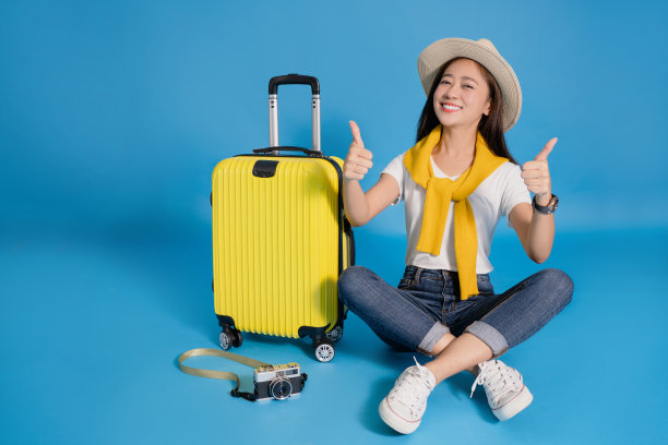 快乐的年轻亚洲女性旅行者坐在一个黄色手提箱在一个蓝色的背景。图片下载
