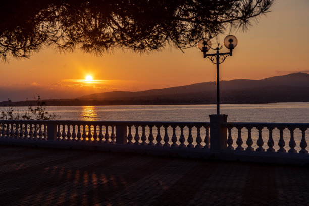 金色的夏日日落。栏杆的剪影。堤岸上栏杆的影子。照明。太阳从山上落下，反射在大海中。松树的树枝在前景。冷静,休息,放松图片下载