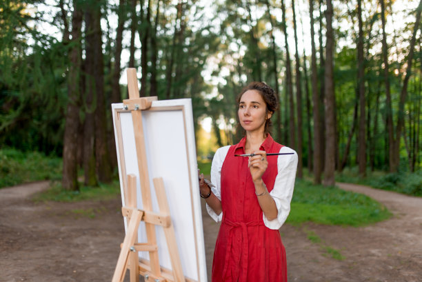 森林中的少女艺术家，情感柔情，轻飘，创造神奇。一名身着红裙的女子，在夏日的自然公园里画画，以树木为背景。画架画照片。创造力的灵感。图片下载