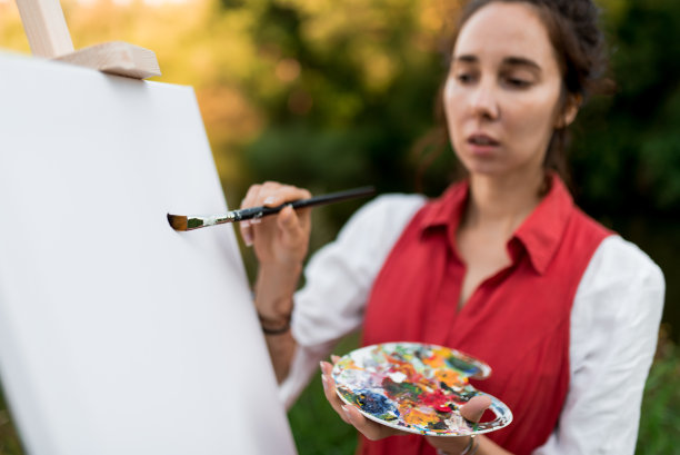 关注刷。绘画的开始。一个穿着红裙子的女人，画着一幅画，在大自然的夏日里，森林里，树木为背景。画架和画笔。灵感和创造力。图片下载