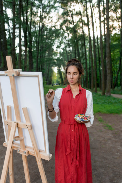 森林里的女画家。一个穿着红色裙子的女人，画了一幅，在夏天的自然公园里，树木为背景。画架和画笔。灵感和创造力。图片下载