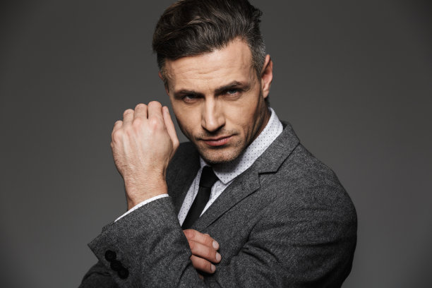 照片特写的商务男子穿着正式的西装，看着相机与严肃的凝视，并展示copyspace手腕，孤立的灰色背景图片下载