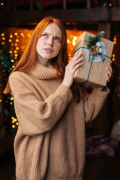 好奇的红发年轻女子摇着包装着圣诞礼物的礼盒。图片下载