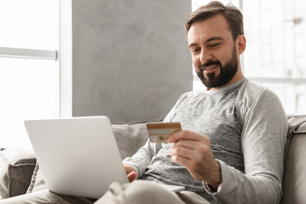 图片是30多岁的黑发男子，穿着便装坐在客厅的沙发上，用信用卡和笔记本电脑进行支付交易图片下载