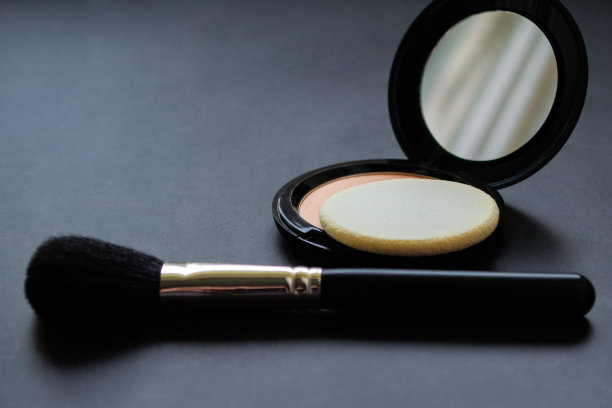 一个黑色的化妆刷，旁边是一个打开的盒子，里面有镜子、粉扑和粉色粉。黑色背景。本空间图片下载