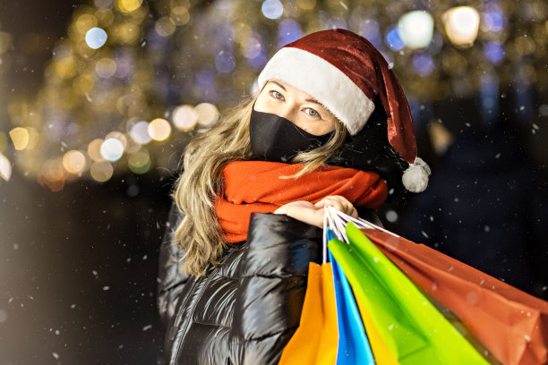 在一家城市商店的橱窗里，一名妇女戴着圣诞帽，戴着黑色医用防护面具，手里拿着装在多色纸袋里的商品。新冠肺炎疫情下的新年购物图片下载