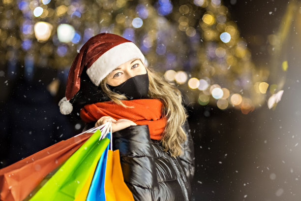 在一家城市商店的橱窗里，一名妇女戴着圣诞帽，戴着黑色医用防护面具，手里拿着装在多色纸袋里的商品。新冠肺炎疫情下的新年购物图片下载