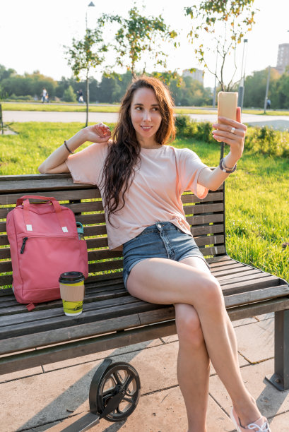 夏天的女人坐在街上的长凳上。照片到手机，在线申请，视频通话记录到智能手机。牛仔短裤和粉色t恤，摩托车和包。图片下载