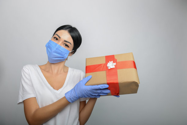 年轻的女孩戴着防护口罩和医用手套，手里拿着一个礼品包。圣诞节前惊喜卫生礼品包。图片下载