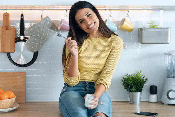 漂亮的年轻女子看着相机，同时吃酸奶坐在家里的厨房。图片下载