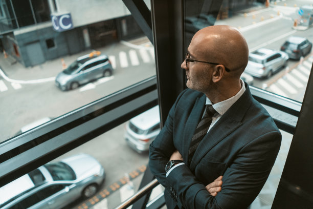 角度视图。穿着西装、戴着眼镜的秃顶商人，在繁忙的街景中望着窗外或玻璃电梯。沉思的商人望着窗外图片下载