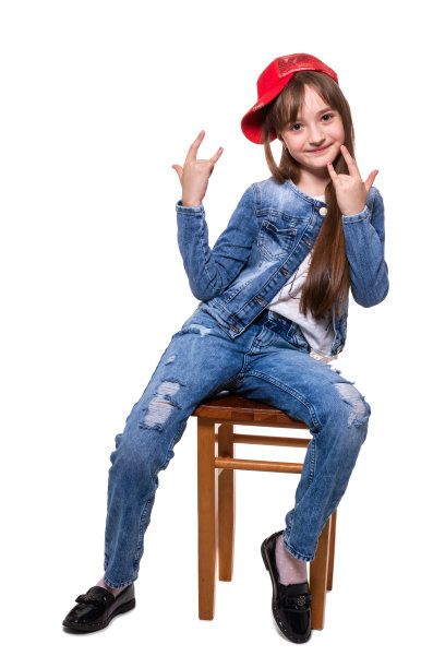 小活泼的女孩穿着牛仔裤套装，戴着红帽子，坐在凳子上摆姿势。在白色背景上的隔离图片下载