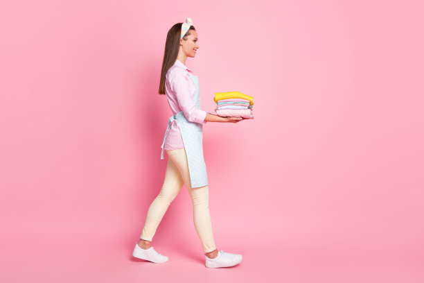 丰满的身材侧面照片，积极开朗的女孩，女佣，抱整齐的堆堆衣服，走，带洗衣，穿裤子，裤子孤立在柔和的颜色背景图片下载