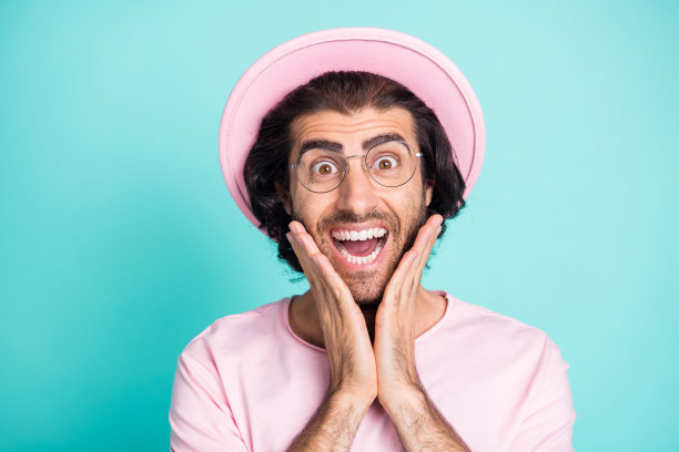 照片肖像惊讶的快乐的男人戴眼镜，粉红色的休闲衣服触摸脸颊孤立在充满活力的蓝绿色背景图片下载