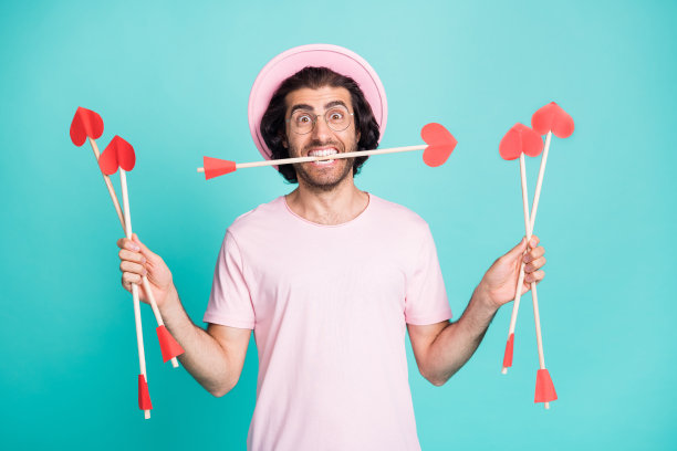 惊讶的潮人男子持箭头穿粉红色帽子眼镜t恤孤立在蓝绿色背景的照片图片下载