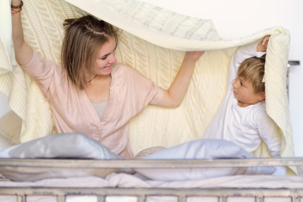 妈妈和孩子在卧室的床上玩得开心，在毯子下面玩。年轻的妈妈带着年幼的儿子一起在周末的床上玩耍，慵懒的早晨，温馨舒适的景象图片下载